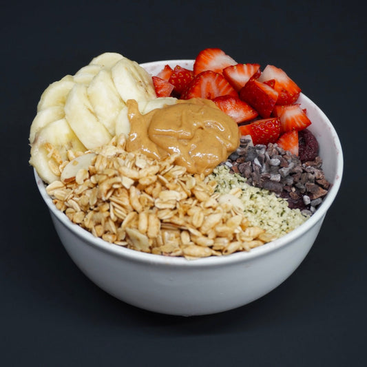 A white bowl filled with a granola smoothie or fruit bowl, topped with sliced bananas, chopped strawberries, peanut butter, oats, hemp seeds, chocolate chips, and dark chocolate pieces, set against a dark background.